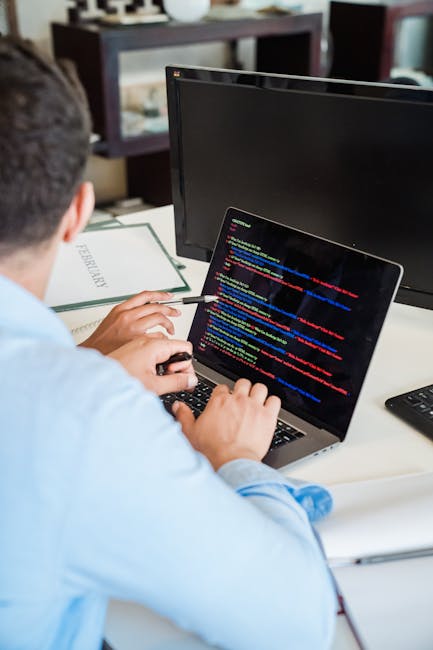 A person working at a desk with a laptop displaying colorful lines of code, surrounded by a monitor, a white paper titled 'FERNARLY' in the background, and a remote control on the desk in a well-lit r