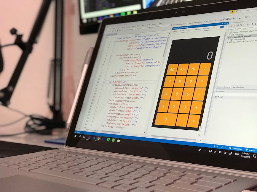 A person working at a desk with a laptop displaying colorful lines of code, surrounded by a monitor, a white paper titled 'FERNARLY' in the background, and a remote control on the desk in a well-lit r