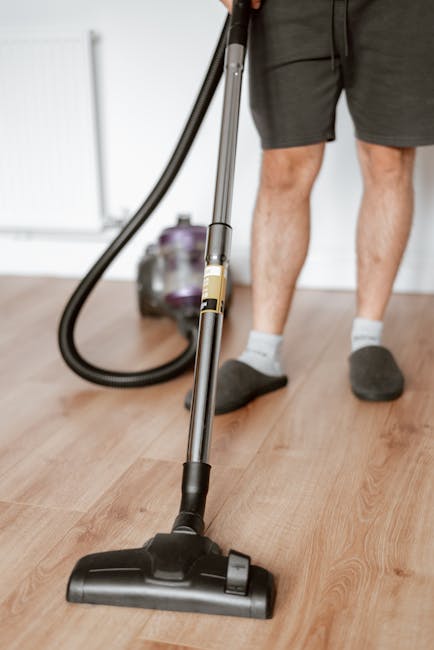 A person dressed in beige overalls is engaged in commercial cleaning on a carpeted floor, using a yellow professional vacuum cleaner with a black hose attachment. The setting appears to be a modern co